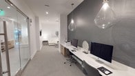 This single-floor space showcases a refined, modern aesthetic defined by a carefully curated palette of whites, grays, and natural wood accents. The workstation area is a standout feature, offering a long, clean-lined white desk equipped with multiple computer terminals, ideal for productivity. Above the desk, teardrop-shaped glass pendant lights hang elegantly, casting a warm, focused glow over the workspace. A bold charcoal herringbone textured accent wall anchors this area, providing a striking contrast against the crisp white walls and light gray carpeted flooring. Recessed ceiling lighting supplements the pendants, ensuring the space remains well-lit throughout. The corridor flows seamlessly into a lounge area, where a neutral-toned seating arrangement and soft ambient lighting create a more relaxed atmosphere. Framed wall art and subtle decorative accents add personality without overwhelming the clean design. The overall ambiance is professional yet inviting, blending functional workspaces with comfortable areas, making this space well-suited for both focused work and casual interaction.