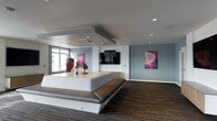 This stylish single-floor space offers a sleek, contemporary aesthetic designed for comfort and social interaction. The open-plan layout is anchored by a large, low-profile platform seating unit with wraparound upholstered benches in a rich brown leather, serving as a central gathering focal point. A wall-mounted flat-screen television is neatly integrated into a white media console, making the space well-suited for presentations or entertainment. The flooring features a textured dark carpet with a subtle pattern, adding warmth and depth to the overall design. Recessed lighting is thoughtfully embedded throughout the ceiling, with a distinctive dropped ceiling detail above the seating area housing an integrated HVAC unit, contributing to a clean and architectural look. The cool grey-blue tones of the walls create a calm, modern atmosphere, further elevated by two bold abstract artworks in pink and magenta hues that add striking visual contrast and personality. A window along one wall introduces natural light, brightening the space and enhancing its open feel. The overall ambiance is professional yet inviting, making it well-suited as a lounge, meeting area, or communal gathering space.