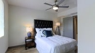 This single-floor space offers a refined and comfortable living environment highlighted by thoughtful design and quality finishes. The bedroom serves as a standout feature, centered around a striking queen-sized bed with a bold, tufted black leather headboard that anchors the room with a sense of sophistication. Crisp white bedding layered with decorative pillows in navy and green adds a touch of color and personality to the space. Warm ambient lighting is provided by two coordinating table lamps — one with a ceramic base and another on a distinctive tripod floor lamp stand — casting a soft, inviting glow throughout the room. A modern ceiling fan with a brushed nickel finish is mounted overhead, offering both style and practical air circulation. The light wood-toned flooring adds warmth and continuity to the space, complementing the neutral, soft gray walls that create a calm and versatile backdrop. Subtle architectural details, including a smooth ceiling and clean door trim, contribute to the overall polished aesthetic. The layout feels well-organized and spacious, making efficient use of the available area while maintaining an uncluttered, comfortable atmosphere suitable for rest and relaxation.