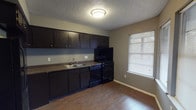 This efficient single-story, 1-bedroom, 1-bathroom unit is thoughtfully designed to make the most of its compact footprint. The layout flows openly between the living area and kitchen, creating a connected and functional living space. The kitchen is well-appointed with dark espresso-toned cabinetry offering generous upper and lower storage, dark countertops, a double-basin sink, and a full suite of black appliances including a refrigerator, range, and dishwasher. A window with blinds brings in natural light, brightening the cooking space. The living area features warm wood-look hard surface flooring that extends throughout the unit, neutral gray walls, and a textured ceiling. Multiple windows dressed with horizontal blinds allow ample daylight into the space, enhancing the open feel. A dedicated accent wall frames the entry door, adding a subtle design touch. The bedroom is accessible through a doorway and continues the same flooring and neutral color palette. The bathroom includes a full tub and shower combination with chrome fixtures, white tile surround, and a small window for ventilation and natural light. Overall, the unit presents a cohesive, practical design suited for comfortable everyday living.