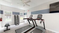 Located at 122 Riverstone Ct in Easley, SC, this fitness center is a thoughtfully designed exercise space that delivers a focused and functional workout environment. The room features a clean, modern aesthetic defined by a bold horizontal stripe of slate blue paint that wraps around the white walls, lending a polished, energetic feel to the interior.

The space is equipped with multiple Star Trac treadmills and an upright stationary bike, providing dedicated cardio options for users. A full rack of dumbbells lines one wall, offering a comprehensive range of free weights for strength training. A large wall-mounted mirror expands the sense of space and allows users to monitor their form during exercise.

Natural light filters in through a side window and a set of glass double doors, which serve as the primary entrance and offer views to the outdoors. A wall-mounted flat-screen television is positioned for easy viewing from the cardio equipment. Ceiling fans throughout aid in air circulation, and a cassette-style HVAC unit in the ceiling ensures climate comfort year-round. A wall-mounted water fountain provides convenient hydration access.

Underfoot, durable carpet tile covers the floor, and crisp white trim and recessed lighting complete the bright, well-maintained atmosphere of this practical community fitness facility.