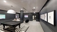This single-floor space is designed as a modern lounge and recreation area, combining style and functionality in a cohesive, polished environment. The interior is anchored by a dark-finished billiards table set atop a bold black-and-white striped area rug, creating a striking focal point upon entry. Adjacent to the billiards area, a comfortable seating arrangement features a light-toned sofa and a sleek accent chair, providing a relaxed gathering space centered around a wall-mounted flat-screen television. The room's design palette leans heavily into a sophisticated dark aesthetic, with charcoal-toned walls and structural columns that lend depth and dimension to the space. Recessed lighting throughout the drop ceiling ensures even, ambient illumination across the room. A series of framed artworks lines the right-hand wall, functioning as a curated gallery that adds a refined, artistic element to the environment. Light-toned carpeting runs throughout, softening the contrast of the darker walls and furnishings. The layout is open and spacious, allowing for easy movement between the various activity zones. The overall ambiance is contemporary and upscale, making the space well-suited for both casual recreation and social gatherings.
