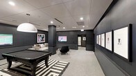 This single-floor space is designed as a modern lounge and recreation area, combining style and functionality in a cohesive, polished environment. The interior is anchored by a dark-finished billiards table set atop a bold black-and-white striped area rug, creating a striking focal point upon entry. Adjacent to the billiards area, a comfortable seating arrangement features a light-toned sofa and a sleek accent chair, providing a relaxed gathering space centered around a wall-mounted flat-screen television. The room's design palette leans heavily into a sophisticated dark aesthetic, with charcoal-toned walls and structural columns that lend depth and dimension to the space. Recessed lighting throughout the drop ceiling ensures even, ambient illumination across the room. A series of framed artworks lines the right-hand wall, functioning as a curated gallery that adds a refined, artistic element to the environment. Light-toned carpeting runs throughout, softening the contrast of the darker walls and furnishings. The layout is open and spacious, allowing for easy movement between the various activity zones. The overall ambiance is contemporary and upscale, making the space well-suited for both casual recreation and social gatherings.