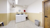 Located at 4440 Beard Ave S in Minneapolis, MN, this shared laundry room is a practical and neatly maintained facility designed for everyday use. The space features a classic mid-century aesthetic, characterized by warm yellow square wall tiles that line the lower half of the walls, complemented by white upper walls and a speckled mosaic tile floor that adds texture and durability underfoot. Two commercial-grade Speed Queen washing machines and matching dryers are positioned along the walls, providing reliable laundry capacity. A wall-mounted folding table offers a convenient surface for sorting and folding laundry, while overhead cabinets provide additional storage. A utility sink sits adjacent to the washers for added functionality. The room is illuminated by flush-mounted ceiling lighting, and a ceiling fan aids in ventilation. A small seating bench offers a place to wait, and a framed artwork on the wall adds a subtle decorative touch. A window along one wall allows for natural light and a view of the surrounding brick exterior. Safety and utility features, including an emergency pull station and utility connections, are also present. The overall ambiance is clean, functional, and straightforward, making it a well-organized shared laundry facility.