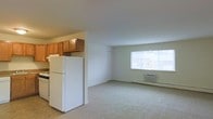 This single-floor space offers a practical and comfortable open-concept layout. The kitchen is equipped with warm honey-toned wood cabinetry, a white refrigerator, and a gas range, providing ample storage and a functional cooking area. The kitchen flows seamlessly into the living area, which features neutral wall-to-wall carpeting that adds warmth underfoot. A large picture window allows generous natural light to fill the living space, creating a bright and welcoming atmosphere. A wall-mounted air conditioning unit and a baseboard heater ensure year-round climate comfort. The neutral color palette throughout — from the light beige walls to the soft carpet — creates a clean, versatile backdrop suitable for a variety of décor styles. The straightforward, single-level layout maximizes usability and accessibility, making it easy to navigate between the kitchen and living areas. Overall, this space combines essential amenities with a simple, functional design that prioritizes comfort and practicality.