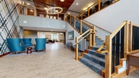 Located at 11001 Bren Rd E in Minnetonka, MN, this thoughtfully designed communal space offers a welcoming and upscale environment characterized by warm natural materials and modern aesthetics. The grand lobby serves as the heart of the space, showcasing soaring double-height ceilings, a dramatic staircase with blue carpeted treads, rich wood handrails, and sleek black metal balusters. A statement circular pendant light anchors the ceiling, while light wood-look tile flooring runs consistently throughout, creating a cohesive and airy feel.

The lounge area features floor-to-ceiling windows that flood the space with natural light, a linear fireplace set against a striking wood-clad accent wall, and a curated arrangement of mid-century modern seating in deep greens, blues, and earth tones. A built-in shelving unit with teal cabinetry adds both function and visual interest to a cozy reading nook.

The social hub includes a bar-height communal counter with decorative geometric paneling, pendant lighting, and a fully equipped kitchenette with dark tile backsplash and stainless steel appliances. A dedicated dining area with bold patterned chairs and teal accent walls pays homage to the local Lake Minnetonka surroundings through vibrant artwork. Corridor areas feature eye-catching geometric wallcoverings, completing the polished and distinctive design throughout.