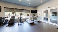 Located at 200 S Mallory St in Hampton, Virginia, this well-appointed fitness center comprises two distinct exercise rooms designed to support a comprehensive workout experience. The main cardio and strength room features durable rubber flooring, recessed lighting, and large windows that flood the space with natural light while offering outdoor views. It is equipped with treadmills, elliptical machines, a stationary bike, a cable/functional trainer, and a dumbbell rack, along with a wall-mounted flat-screen TV for entertainment during workouts. Full-length mirrors line one wall, enhancing the sense of openness throughout the room. A set of glass double doors provides direct access to an outdoor patio area, further connecting the interior to the surrounding environment. The secondary studio room offers a more versatile, open-floor setting finished with the same rubber flooring and neutral gray walls. It is outfitted with stability balls, medicine balls, resistance bands, spin bikes, and a wall-mounted TV, making it well-suited for group fitness, stretching, or individual training sessions. Colorful fitness-themed artwork decorates the walls of both rooms, contributing to an energetic and motivating atmosphere throughout the facility.