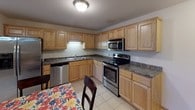This single-floor residence offers a practical and comfortable layout with multiple bedrooms, two bathrooms, and a fully equipped kitchen. The kitchen is a functional centerpiece, featuring warm oak cabinetry with ample storage, dark granite-style countertops, stainless steel appliances including a range, over-the-range microwave, dishwasher, and refrigerator, all set on light ceramic tile flooring. The bedrooms are carpeted and finished with neutral warm tones, each benefiting from natural light through wood-framed windows fitted with blinds. At least one bedroom includes a walk-in closet with built-in shelving and hanging rods, while another features a standard closet with an oak door. The bathrooms are finished with light ceramic tile flooring, oak vanities with white countertops, and well-lit mirrors. One bathroom includes a sliding glass door shower enclosure. Throughout the space, consistent design elements tie the rooms together, including light oak interior doors, neutral beige walls, and textured ceilings. The floor plan, as seen from above, reveals an efficient and cohesive layout that connects the kitchen, living, and sleeping areas in a straightforward, accessible arrangement.