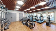 Located at 3223 Timber Mill Pkwy in Castle Rock, Colorado, this fitness center is a thoughtfully designed exercise space that combines modern aesthetics with comprehensive workout functionality. The room's most distinctive architectural feature is its eye-catching slatted wood ceiling, where warm-toned wooden slats run in parallel formation and are punctuated by large circular flush-mounted light fixtures that provide bright, even illumination throughout the space.

The flooring consists of a parquet-style tile in warm earth tones, contributing to the inviting atmosphere of the room. A row of windows along one wall allows natural light to fill the space and offers views to the outdoors, while full-length mirrors on adjacent walls visually expand the room and allow users to monitor their form during workouts.

The exercise floor is comprehensively equipped with a variety of cardio machines, including multiple treadmills and a stationary bike, as well as strength-training equipment such as a cable machine, leg press, and adjustable weight bench. A dedicated free weights area features a full dumbbell rack alongside additional benches and stability balls. Storage shelving provides convenient organization for accessories. Gold-toned wall sconces add a refined decorative touch, rounding out this modern and well-appointed fitness facility.
