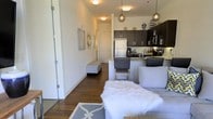 This single-floor space showcases a thoughtfully designed open-concept layout that connects the living area and kitchen in one cohesive environment. Rich hardwood flooring runs throughout, adding warmth and continuity to the space. The kitchen is appointed with dark espresso cabinetry, a white refrigerator, stainless steel appliances, and a kitchen island or peninsula with seating, making it both functional and visually striking. Pendant lighting with globe and metallic fixtures hangs above the kitchen area, providing focused illumination while contributing to the modern aesthetic. The living area is comfortably proportioned, featuring a light-toned sofa, a plush white faux-fur accent piece, and a patterned area rug that adds texture to the space. Wall art and a decorative mirror adorn the walls, lending a curated, stylish feel. A hallway extends toward what appears to be additional rooms or an entryway, flanked by white walls and accented with recessed lighting. The overall ambiance is contemporary and polished, with a neutral color palette of whites and grays balanced by the depth of the dark cabinetry and warm wood flooring.