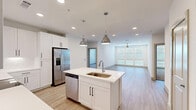 Located at 17501 Dallas Parkway in Dallas, TX, this thoughtfully designed two-bedroom, two-bathroom residence offers a well-appointed single-floor layout with clean, contemporary finishes throughout. The open-concept kitchen and living area serves as the heart of the home, featuring a central island with a built-in sink, white shaker cabinetry extending to the ceiling, stainless steel appliances including a French door refrigerator, microwave, and range, and striking pendant lights overhead. Light wood-style flooring flows through the main living areas, complemented by recessed lighting and a ceiling fan that enhance both function and style. A wall of large windows floods the living space with natural light and offers outdoor views. The two bedrooms are carpeted for comfort, each equipped with a ceiling fan and generous closet space. The primary bedroom features a walk-in closet with built-in shelving and hanging rods. Both bathrooms are finished in white with wood-look flooring, illuminated vanity mirrors, white cabinetry with matte black hardware, and white quartz countertops. The primary bath also includes a soaking tub. This residence combines modern design with practical living in a bright, welcoming environment.