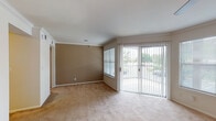 This single-story home offers a well-organized layout with distinct living areas, each finished with practical, durable materials. The main living room is a generously sized space featuring warm beige carpeting, crown molding, and a striking stone-surround fireplace with a classic mantel. A ceiling fan provides year-round comfort, while a large window allows abundant natural light. Sliding glass doors open directly to an outdoor patio area, providing views of surrounding greenery.

An adjoining area, visible through an open passageway, extends the living space and connects to the rest of the home. Crown molding and consistent neutral wall tones carry throughout, contributing to a cohesive aesthetic. A recessed niche adds an architectural detail to the shared wall between spaces.

The galley-style kitchen is efficiently designed with light wood cabinetry lining both sides, offering ample upper and lower storage. White appliances, including a range and dishwasher, complement the light countertops, while tile flooring adds easy maintenance. A set of double doors provides additional pantry or utility storage.

The bedroom features plush carpeting, a ceiling fan with light fixture, and a window with outdoor views. The bathroom includes a single-sink vanity with wood cabinetry, a large mirror, and a full tub-shower combination with white tile surround and overhead cabinetry for added storage.