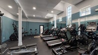Located at 2201 Greenville Blvd in Greenville, NC, this single-floor fitness center is designed to support a comprehensive workout experience. The space features a bold two-tone color scheme, with deep teal accent walls contrasting against crisp white upper walls, creating an energizing atmosphere throughout. Recessed lighting and a row of tall, multi-pane windows along the perimeter flood the facility with natural light, while large wall-mounted mirrors amplify the sense of openness and allow members to monitor their form.

The cardio zone is well-equipped with multiple treadmills, ellipticals, and stationary bikes arranged in an organized row for easy access. Adjacent to the cardio area, a dedicated free weights section includes a rack of dumbbells, adjustable benches, and a cable machine to accommodate a range of strength training routines. Structural columns integrated into the layout divide the space naturally without interrupting flow.

A separate functional training area provides a distinct workout environment, featuring a multi-station cable/pulley system, a leg press machine, a heavy bag with an MMA stand, TRX suspension straps, and a barbell. A bold motivational mural anchors this section, reinforcing the fitness-focused atmosphere. The dark rubber flooring throughout the facility ensures durability and safety, making this a fully equipped destination for diverse fitness goals.
