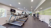 This fitness center offers a comprehensive workout environment thoughtfully designed to inspire and motivate. The elongated layout is anchored by a row of Matrix treadmills and elliptical machines, providing ample cardio options for users of all fitness levels. Recessed lighting throughout the ceiling ensures the space is consistently well-lit, creating a clean and energizing atmosphere.

The walls serve as a standout design feature, adorned with bold fitness-themed motivational words and phrases — such as "inspire," "focus," "invigorate," and "conquer" — rendered in a vibrant lime green palette against crisp white walls. These graphic accents contribute to an upbeat and encouraging ambiance throughout the facility.

A dedicated strength training section includes a cable and weight machine setup, a dumbbell rack, and additional resistance equipment, catering to a full range of strength workouts. A separate area houses recumbent bikes and stability balls on a rack, ideal for low-impact and functional training. Large wall-to-wall mirrors further expand the sense of space and allow users to monitor their form.

The durable speckled rubber flooring provides safety and comfort underfoot, while a small refreshment station with a water cooler adds a practical convenience. Windows allow for a touch of natural light, complementing the recessed overhead lighting for a bright and welcoming workout environment.