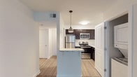 Cardinal is a thoughtfully designed single-floor residence that combines modern finishes with functional living spaces. The kitchen serves as a central highlight, featuring dark espresso cabinetry, light granite-style countertops, and a full suite of stainless steel appliances including a refrigerator, range, microwave, and dishwasher. A breakfast bar with pendant lighting provides a casual dining option and creates a natural transition between the kitchen and the living area. Wood-style flooring runs throughout the kitchen, adding warmth and easy maintenance to the space. The living area is carpeted and generously proportioned, bathed in natural light from a large sliding glass door that opens to an outdoor view. French-style interior doors connect the living space to an additional room, offering flexibility for various uses. Convenient in-unit laundry is tucked just off the kitchen, providing added practicality. The bathroom is clean and modern, appointed with a granite-style vanity countertop, dark cabinetry, a well-lit mirror, and a full tub-shower combination. Crisp white walls and consistent finishes throughout the residence create a cohesive, move-in-ready atmosphere that is both functional and inviting.