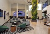 This contemporary commercial lobby makes a striking first impression with its soaring double-height ceilings that flood the space with an open, airy ambiance. Large-format light stone tile flooring extends throughout, lending a clean and cohesive aesthetic underfoot. A dramatic living green wall anchors one side of the space, introducing a vibrant natural element that contrasts beautifully with the crisp white walls and modern finishes.

Steel-framed glass partitions divide the lobby from adjacent office and work areas, maintaining visual connectivity while defining functional zones. A bold staircase with dark wood treads and black metal railings rises prominently, leading to an upper-level mezzanine overlooking the lobby below. Seating areas are thoughtfully arranged, featuring a mix of gray sofas, green accent chairs, and patterned area rugs that add warmth and texture.

A central round table with low cork stools creates an informal gathering point, while wall-mounted displays provide informational content throughout. Potted plants and sculptural floor lamps contribute to the layered, curated atmosphere. Natural light enters through clerestory windows and glass entry doors, further enhancing the bright, welcoming character of this well-designed professional environment.