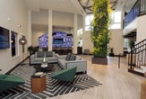 This contemporary commercial lobby makes a striking first impression with its soaring double-height ceilings that flood the space with an open, airy ambiance. Large-format light stone tile flooring extends throughout, lending a clean and cohesive aesthetic underfoot. A dramatic living green wall anchors one side of the space, introducing a vibrant natural element that contrasts beautifully with the crisp white walls and modern finishes.

Steel-framed glass partitions divide the lobby from adjacent office and work areas, maintaining visual connectivity while defining functional zones. A bold staircase with dark wood treads and black metal railings rises prominently, leading to an upper-level mezzanine overlooking the lobby below. Seating areas are thoughtfully arranged, featuring a mix of gray sofas, green accent chairs, and patterned area rugs that add warmth and texture.

A central round table with low cork stools creates an informal gathering point, while wall-mounted displays provide informational content throughout. Potted plants and sculptural floor lamps contribute to the layered, curated atmosphere. Natural light enters through clerestory windows and glass entry doors, further enhancing the bright, welcoming character of this well-designed professional environment.