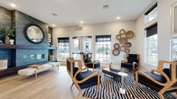 This single-floor space functions as a well-appointed clubhouse or leasing center, thoughtfully designed to blend contemporary aesthetics with welcoming comfort. The layout is open and expansive, with wide-plank light hardwood flooring flowing seamlessly throughout every area, contributing to a bright, cohesive atmosphere.

The reception and leasing area is defined by a sleek workstation enclosed in floor-to-ceiling glass panels with dark metal framing, creating a professional yet transparent workspace. Velvet upholstered chairs in muted gray tones line the front of the desk, while tall windows along the perimeter flood the room with natural light. Sculptural wall art panels and large framed works add a gallery-quality presence to this area.

Adjacent lounge spaces offer a more relaxed environment. One features a striking dark stacked-stone fireplace with a round beaded mirror as a focal point, paired with wood-framed accent chairs upholstered in deep navy fabric and arranged over a bold geometric area rug. French doors provide direct outdoor views and natural light.

A separate seating nook showcases a dramatic navy accent wall adorned with a large-scale metallic sculptural artwork, complemented by a cream sofa, plaid accent pillows, and a sculptural dark wood coffee table. Woven basket wall décor and recessed lighting throughout complete the refined, curated ambiance.