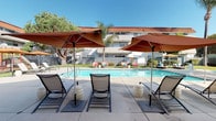 Located at 9128 Burke St in Pico Rivera, California, this single-level community amenity space is designed to offer residents a welcoming outdoor retreat with a range of shared facilities. The centerpiece is a generously sized outdoor swimming pool surrounded by a spacious concrete deck, furnished with multiple lounge chairs and large terracotta-toned market umbrellas that provide ample shade for relaxation under the Southern California sun. Mature palm trees and lush greenery frame the pool area, complementing the Spanish-influenced architecture of the surrounding buildings, which feature classic red tile roofing. Adjacent to the pool area is a covered outdoor lounge space with a clean, mid-century-inspired aesthetic. This shaded pavilion features a wood-paneled ceiling supported by concrete columns and opens to views of swaying palm trees and the surrounding grounds. The space is furnished with comfortable seating, including a vibrant red sofa, and is equipped with vending machines and an outdoor bar counter with seating, making it a convenient gathering spot. A transitional passageway connects the covered area to the broader grounds, featuring restroom access and open sightlines to the well-maintained green spaces beyond. The overall ambiance is relaxed, functional, and suited to communal outdoor living.