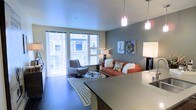 This single-floor space showcases a thoughtfully designed open-concept layout that seamlessly connects the kitchen and living areas. The kitchen features sleek countertops and pendant lighting, with a generous island or bar-height counter that serves as both a functional workspace and a visual divider between the cooking and living zones. Warm pendant lights hang above, casting a welcoming glow throughout.

The living area is anchored by a vibrant orange sofa accented with decorative pillows, complemented by a modern swivel accent chair and a patterned area rug that adds texture to the dark hardwood-style flooring. A glass-topped coffee table and carefully selected wall art contribute to the space's contemporary character.

Large floor-to-ceiling windows flood the room with natural light and offer views of the surrounding building exterior, creating a bright and airy atmosphere. Neutral tones on the walls are offset by a bold accent wall in a deep teal-gray, adding depth and visual interest to the overall design. The space strikes a balance between modern sophistication and comfortable livability, making it an inviting environment well-suited for both relaxing and entertaining.