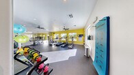 Located at 10200 S Giles St in Las Vegas, NV, this fitness center offers a bright and energetic exercise space designed to support a full range of workout routines. The single-room facility is characterized by bold yellow accent walls that energize the environment, complemented by white ceilings and durable dark rubber flooring suited for high-activity use. Large windows line the walls, flooding the space with natural light and providing views of the surrounding outdoor area, including what appears to be a pool and landscaped grounds. Dual ceiling fans enhance air circulation throughout the room, contributing to a comfortable workout environment. The exercise room is comprehensively equipped with multiple treadmills, elliptical machines, and a stationary recumbent bike for cardio training. A dedicated TRX Training Zone is mounted on the wall, accompanied by instructional charts. Free weight offerings include a full dumbbell rack and an assortment of colorful kettlebells. Stability balls, medicine balls, balance discs, and foam rollers round out the functional fitness equipment. Floor-to-ceiling mirrors on one wall amplify the sense of space and allow users to monitor their form. A Smart Mat exercise guide is also displayed for reference. The overall ambiance is vibrant, well-lit, and purposefully arranged to maximize usability.