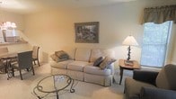 This single-floor space features a living area designed with a traditional and understated aesthetic. The room is finished with wall-to-wall carpeting in a soft, neutral tone that contributes to its warm and comfortable atmosphere. The walls are painted in a light, creamy hue, creating a bright and airy feel throughout the space.

The living area is furnished with a generously sized, upholstered sofa in a pale, textured fabric, accented with decorative throw pillows in muted earth tones. A glass-topped coffee table with an ornate wrought-iron base serves as a central focal point, providing both function and visual interest. A wooden side table positioned near the sofa supports a classic table lamp with a warm-toned shade, adding soft, ambient lighting to the room.

A framed landscape painting adorns the wall above the sofa, offering a touch of classic decorative style. The overall arrangement of the furniture encourages conversation and relaxation. The space maintains a cohesive, traditional design language with its coordinated furnishings, neutral palette, and carefully selected décor elements, making it a comfortable and inviting living environment.