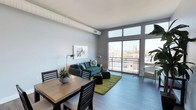 This contemporary single-floor urban residence offers a thoughtfully designed open-concept layout that seamlessly blends living and dining areas. The space is anchored by expansive floor-to-ceiling windows that flood the interior with abundant natural light while framing stunning views of the city skyline and surrounding urban landscape. A sliding glass door provides direct access to a private balcony, extending the living space outdoors and offering an elevated vantage point over the city below. The living area features stylish modern furnishings, including a teal sofa accented with decorative pillows and a vibrant green area rug that adds a pop of color against the warm hardwood flooring. The dining area is defined by a wood-top table paired with dark wooden chairs, creating a welcoming space for meals and gatherings. Exposed ductwork along the ceiling adds an industrial design element that complements the space's modern urban aesthetic. Neutral gray walls provide a clean, versatile backdrop throughout, while carefully placed greenery and artwork contribute to a warm and inviting ambiance. The overall design strikes a balance between contemporary style and comfortable, functional living.