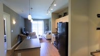 This single-floor space is designed with an open-concept layout that seamlessly connects the kitchen and living area, creating a cohesive and functional environment. The kitchen is outfitted with dark, stainless-finish appliances, including a black refrigerator and range, paired with light-toned cabinetry that provides ample storage. A peninsula countertop with a double sink runs along one side, offering both workspace and a natural visual divider between the kitchen and the adjoining living area. Pendant lights hang above the peninsula, complementing a track lighting system overhead that runs the length of the space, ensuring thorough and adjustable illumination throughout.

Warm wood-look flooring extends consistently across the entire floor plan, contributing to a unified and inviting aesthetic. The living area is anchored by a comfortable seating arrangement set atop a decorative area rug. At the far end of the space, a full-height sliding glass door floods the interior with natural light and provides an open, airy quality. Potted plants are placed thoughtfully on top of the cabinetry and near the entrance, adding natural accents that soften the modern design. The overall ambiance is contemporary, warm, and practical.