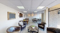 Located at 779 Eves Dr in Hillsborough, NJ, this well-appointed community lounge and gathering space offers a comfortable and functional environment designed for socializing, dining, and small meetings. The single-floor layout is thoughtfully organized into distinct zones, including a relaxed lounge area furnished with rich leather club chairs and a marble-topped coffee table, and multiple dining areas with warm wood tables and modern metal chairs. A wall-mounted flat-screen TV anchors one end of the main space, while decorative wall sconces and inspirational signage add a welcoming touch throughout. The drop-panel ceiling with recessed and flush lighting ensures the space is consistently and evenly lit. A dedicated conference nook features a large round clock as a focal point, making it ideal for focused meetings or group sessions. Large floor-to-ceiling windows and glass sliding doors flood the interior with natural light and provide picturesque views of the surrounding mature evergreen trees. The sliding doors open to an outdoor patio area, seamlessly blending indoor comfort with outdoor access. Patterned carpeting in soft neutral tones ties the space together, creating a cohesive and inviting atmosphere suitable for a variety of community activities.