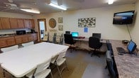 This single-floor multipurpose room is designed to serve a variety of communal functions, combining practical amenities with a welcoming atmosphere. The space is anchored by a large white folding table surrounded by folding chairs, providing ample room for group gatherings, meetings, or shared activities. Overhead, ceiling fans and fluorescent light fixtures ensure the room stays well-lit and comfortable throughout the day.

Along one wall, a kitchenette offers wooden cabinetry, a sink, a microwave, and a coffee maker, making the space well-suited for events or everyday use. A door near the kitchenette leads to an adjacent area, adding to the overall flow of the space.

On the opposite side of the room, a dedicated computer workstation area features multiple desktop computers with monitors, office chairs, and a long shared desk — providing a functional workspace for residents or visitors. Additional folding chairs are stored neatly nearby for flexible use.

The walls are decorated with tasteful wall art, including a circular decorative piece and floral panels, giving the room a warm and lived-in feel. The floor features a mixed-tile and carpet design, subtly defining different functional zones within the room. Overall, this space balances practicality with comfort, serving effectively as a shared community hub.