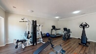 Located at 1806 Maplewood Ln in Greensboro, North Carolina, this single-floor fitness facility is purpose-built to support a comprehensive workout experience. The main exercise room is generously sized, finished with rich, dark hardwood-style flooring and neutral gray walls that create a clean, focused environment. A textured ceiling with flush-mount lighting provides consistent illumination throughout the space, while crown molding adds a refined architectural touch.

The room is well-appointed with a diverse selection of fitness equipment, including a multi-station cable and weight machine, a treadmill, a recumbent bike, an upright stationary bike, adjustable weight benches, resistance and rowing equipment, and stability balls — offering options for both cardiovascular and strength training. A wall-mounted TV bracket is positioned for convenient viewing during workouts, and a full-length mirror supports proper form and technique.

Natural light filters in through multiple windows, including a large window on one wall, enhancing the airy feel of the space. A set of glass French doors provides direct access to the outdoors, contributing additional brightness and convenient entry and exit. Safety features include a prominently placed fire extinguisher near the exit. A small counter area near the entry door offers a practical space for personal items. The overall layout is open and functional, designed to accommodate a variety of fitness routines efficiently.