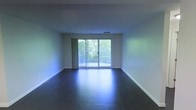 This single-floor space is designed with a clean, minimalist aesthetic that emphasizes openness and natural light. The main living area features dark hardwood flooring that contrasts elegantly with the bright white walls and white baseboards, creating a modern and polished look throughout. The room is generously proportioned, offering a spacious and airy feel well-suited for a variety of furniture arrangements.

A prominent sliding glass door serves as the focal point of the main living area, flanked by sheer curtains that diffuse natural light softly into the room. The door opens directly onto a private balcony enclosed by a sleek metal railing, which overlooks lush, mature tree canopy — providing a tranquil and verdant outdoor view. This connection to the outdoors enhances the overall ambiance, lending the interior a bright and refreshing atmosphere even when viewed from within.

The ceiling is smooth and uniform, fitted with a central HVAC vent, contributing to the space's clean and uncluttered character. The neutral color palette throughout the interior allows for versatile interior design possibilities, making it a highly adaptable living environment.
