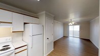 This single-floor space offers a practical and cohesive layout, with the kitchen and living area sharing an open sightline that creates a sense of spaciousness. The kitchen is outfitted with white cabinetry extending to the ceiling, providing ample storage, and includes a full-size white refrigerator. A built-in cabinet with a decorative ventilation panel adds a subtle architectural detail near the transition between the kitchen and living areas. Crown molding runs consistently throughout, lending a clean, finished look to the interior. The flooring throughout is light-toned hardwood, contributing warmth and continuity across the space. The living area is anchored by a ceiling fan with a light fixture, offering both comfort and illumination. A sliding glass door at the far end of the living space allows natural light to filter in and provides a visual connection to the outdoors. The walls are painted in a neutral tone, complementing the bright, airy feel of the space. Overall, the layout is straightforward and functional, with design details that add character without overwhelming the understated aesthetic.