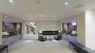 This elegantly designed single-floor space presents a refined, contemporary aesthetic that blends comfort with modern sophistication. The open-plan layout is anchored by a sleek, linear gas fireplace set into a clean white feature wall, creating a warm and inviting focal point for the lounge area. Flanking the fireplace are two stylish high-back accent chairs paired with a round glass side table, offering an intimate seating arrangement ideal for conversation or relaxation. A striking decorative installation of overlapping metallic disc art adorns the adjacent wall, adding texture, warmth, and visual interest to the neutral palette. Recessed lighting throughout the smooth white ceiling provides bright, even illumination that enhances the airy feel of the space. The flooring transitions between polished light-toned surfaces and a boldly patterned area rug that defines the lounge zone and introduces graphic energy underfoot. In the background, an additional dining or lounge area is visible, furnished with chairs and pendant lighting, suggesting a multi-functional layout suited for both social gatherings and quiet retreats. The overall ambiance is calm, curated, and professional — consistent with an upscale hospitality or shared amenity environment.