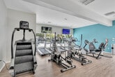 Located at 650 Ebbcreek Dr in Corona, CA, this fitness center is a thoughtfully designed, single-floor facility comprising two dedicated exercise rooms that together offer a comprehensive workout experience. The space is distinguished by its soaring vaulted ceilings, skylights, and track lighting, which flood the interiors with natural and artificial light, creating an open and energizing atmosphere. Light hardwood-style flooring runs throughout both rooms, contributing to a clean, modern aesthetic. The first exercise room is oriented toward cardio and strength training, featuring a row of treadmills, ellipticals, and stationary bikes alongside cable and weight machines. Large windows frame outdoor views of the surrounding landscaping and pool area, while a wall-mounted flat-screen TV keeps workouts engaging. A vibrant abstract mural adds an artistic focal point to one corner. The second exercise room offers a more functional training environment, equipped with a power rack, kettlebell storage, dumbbells of varying weights, and additional cable machines. Teal accent walls paired with floor-to-ceiling mirrors amplify the sense of space. Glass doors open directly to the exterior, enhancing ventilation and connectivity to the outdoors. Together, the two rooms provide a well-rounded, inviting facility suited for a wide range of fitness activities.