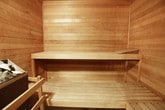 This single-floor wellness facility is designed around two core areas: a traditional sauna room and an adjoining shower space. The sauna room is fully clad in horizontal wood planking across all walls, the ceiling, and the floor, creating a warm, enveloping aesthetic consistent with classic Scandinavian sauna design. A central rock heater filled with natural stones serves as the focal point of the sauna, providing the heat source for steam sessions. Slatted wooden benches line the interior, offering seating and reclining space for users. Instructional signage is mounted on the walls, including notices regarding proper sauna use and heater care. The wood tones range from lighter, naturally finished planks on the benches to richer, darker stained panels on the walls, adding visual depth to the space. Adjacent to the sauna, a separate shower area features a tiled floor, frosted glass shower enclosure panels, and clean white walls, providing a functional and hygienic space for rinsing. A dark-framed doorway connects the two areas, creating a clear transition between the dry heat environment and the wet shower zone. Together, the two spaces form a cohesive and practical wellness retreat.