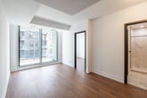 This well-designed one-bedroom, one-bathroom unit offers an efficient layout across a single floor, making effective use of its compact footprint. The living area is characterized by warm hardwood flooring and crisp white walls, creating a clean, modern aesthetic throughout. Large floor-to-ceiling glass panels and sliding doors dominate the main living space, flooding the interior with natural light and providing views of the surrounding urban building facade.

The bedroom is a comfortably sized private retreat, also featuring hardwood flooring and large windows that continue the unit's bright, airy character. White walls and simple finishes keep the space feeling open and uncluttered.

The kitchen area, visible in the unit's layout, is equipped with stainless steel appliances — including a refrigerator and range — set against white cabinetry and light countertops, providing practical functionality within the compact space.

The bathroom is a standout feature, showcasing floor-to-ceiling marble-effect porcelain tile walls and a mosaic tile shower floor. A frameless glass shower enclosure paired with matte black fixtures, including a wall-mounted showerhead and controls, adds a refined, contemporary touch. The overall design of the unit balances modern finishes with practical, efficient use of space.