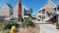 This single-story mail room and common area serves as a central hub for a multi-building residential complex. The exterior of the community showcases a classic design with angled rooflines, neutral lap siding, and private balconies on upper-level units, all set against a backdrop of mature palm trees, pines, and drought-tolerant landscaping that thrives under abundant natural sunlight.

The covered mail area is a practical, open-air structure with a concrete floor and a low-profile ceiling that provides shade while maintaining an airy feel. The space is equipped with an extensive bank of individual resident mailboxes arranged in neat rows, each unit clearly numbered for easy identification. Alongside the standard mailboxes, a set of secure parcel lockers accommodates package deliveries. A wall-mounted bulletin board offers a space for community notices and announcements.

The walkways connecting this area to the surrounding buildings are wide and well-maintained, lined with low-growing shrubs and ground cover plantings. Covered carport parking is visible nearby, and the open sky above the complex provides a bright, sun-filled atmosphere. The overall ambiance is functional and community-oriented, offering residents a convenient and accessible central point for daily mail retrieval within a well-kept outdoor setting.