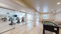 Located at 66 Glenbrook Rd in Stamford, Connecticut, this single-floor amenity space offers a well-appointed fitness and social area designed to support an active lifestyle. The space is finished with light hardwood-style flooring throughout, recessed lighting, and a clean, modern aesthetic defined by white and neutral walls that create a bright, open atmosphere.

The centerpiece of the fitness area is a dedicated spin studio enclosed by full-height glass sliding doors, providing a sleek, studio-gym feel while maintaining visual openness. The cycling room is equipped with multiple stationary spin bikes positioned on protective floor mats, wall-to-wall mirrors that amplify the sense of space, and a wall-mounted flat-screen television. Natural light filters in through large windows, adding to the energizing environment.

Adjacent to the spin studio, a free weights station features a dumbbell rack alongside additional spin bikes, giving residents a versatile strength and cardio training area. A direct exit door with exterior windows provides convenient access and ventilation.

The resident lounge area flows naturally from the fitness space, offering a generous L-shaped sectional sofa in a neutral tone, ideal for relaxing before or after a workout. A foosball table adds a recreational element to the lounge, rounding out the social atmosphere. Together, these spaces form a cohesive, amenity-rich environment suited for both fitness and community engagement.
