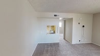 Located at 900 N Country Club Dr in Mesa, Arizona, this single-floor unit offers a functional and straightforward layout throughout. The living area is finished with neutral-toned carpet and features a distinctive pass-through opening to the kitchen, which includes a range and built-in cabinetry, creating a connected feel between the two spaces. A textured popcorn ceiling runs through the main living areas, and centrally positioned HVAC vents provide climate control throughout.

The bedroom is generously proportioned, carpeted, and receives natural light through a sliding window fitted with vertical blinds. An additional room with carpeting and a pass-through kitchen nook with a refrigerator and cooktop provides flexible living or dining options.

The bathroom is well-appointed with a wide vanity featuring a speckled countertop and single-basin sink, a large wall mirror, overhead lighting, a standard toilet, and a shower-tub combination. Dark wood-style vinyl flooring grounds the space with a warm contrast to the light walls.

A dedicated utility area includes upper and lower white cabinetry, a stainless steel utility sink, and a speckled countertop with an electrical outlet, ideal for laundry or storage needs. Throughout the unit, neutral wall tones and consistent finishes create a clean, move-in-ready atmosphere.