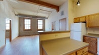 This single-floor space showcases a distinctive loft-style design rooted in industrial heritage. The open-plan layout seamlessly connects the kitchen and living areas, creating a spacious and airy atmosphere enhanced by impressively tall ceilings. Prominent wooden beams run overhead, adding warmth and character to the space, while exposed ductwork reinforces the industrial aesthetic.

The kitchen features laminate countertops with a wraparound breakfast bar that serves as a natural dividing element between the cooking and living zones. A white refrigerator and wood-toned cabinetry complete the functional kitchen setup. The pass-through design of the counter allows for an unobstructed sightline into the expansive living area beyond.

The living area is grounded by warm hardwood flooring and framed by original exposed brick walls, lending the space a rich, textured character. Multiple double-hung windows along the brick wall flood the interior with natural light, contributing to the bright and open feel. A doorway visible in the background hints at additional spaces within the unit. The overall ambiance blends industrial rawness with residential warmth.