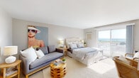 Located at 151 Ocean Rd in Narragansett, Rhode Island, this single-floor coastal retreat offers a thoughtfully designed layout that captures the relaxed elegance of seaside living. The main living and sleeping area is generously proportioned, featuring neutral tones, soft carpeting, and abundant natural light that pours in through a sliding glass door opening to a private balcony with direct ocean views. A tufted upholstered bed, a grey lounge sofa, and a sculptural wood accent table furnish the space with a curated, beach-inspired aesthetic, complemented by a bold pop art portrait that adds a playful artistic touch. A dedicated desk nook with a wooden chair and natural greenery provides a functional corner within the bedroom area. The kitchen is efficiently arranged with white shaker-style cabinetry, light countertops, and a full suite of stainless steel appliances including a refrigerator, range, over-the-range microwave, and dishwasher, all set atop light wood-tone flooring. The bathroom features clean white tile, a glass-enclosed walk-in shower, a white vanity with a mirrored medicine cabinet, and light tile flooring throughout, offering a bright and functional design. Together, the spaces create a cohesive, coastal-contemporary retreat steps from the Rhode Island shoreline.