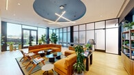 This thoughtfully designed single-floor space offers a compelling blend of modern aesthetics and functional versatility. The main lounge area serves as the centerpiece, featuring a striking circular tray ceiling finished in deep blue and adorned with a sculptural cross-shaped linear pendant light fixture. Warm natural light pours in through floor-to-ceiling windows framed by sleek dark steel mullions, offering open outdoor views and connecting the interior to the surrounding environment. Rich amber velvet sectional sofas and lounge chairs are arranged around a curated collection of dark oval and round coffee tables, creating inviting conversation and relaxation zones. A built-in entertainment wall with a mounted flat-screen TV, open wood shelving, and decorative accents anchors one end of the lounge, while lush indoor plants are thoughtfully placed throughout, adding a vibrant, organic energy to the space. Light oak hardwood flooring runs continuously underfoot, enhancing the warm, welcoming ambiance. A connecting hallway features bold sky-blue accent walls, additional greenery, and stylish shelving, leading to a private meeting room. This intimate room is finished with subtle plaid wallcovering, a round blue table, boucle chairs, and an integrated display screen, making it well-suited for small gatherings or focused work sessions. The space as a whole is polished, design-forward, and conducive to both social interaction and productivity.