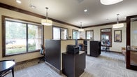 This single-story amenity and leasing space is designed with a cohesive, upscale aesthetic that balances functionality with comfort. The interior is unified by patterned, neutral-toned carpet tiles, warm beige walls, and dark espresso wood trim that frames doorways, windows, and ceiling beams throughout.

The leasing and business area features multiple dark-finished workstations equipped with computers and a printer, all illuminated by pendant drum lights that cast a warm, even glow. Large windows along the perimeter bring in generous natural light and offer views of the surrounding greenery, enhancing the open, welcoming atmosphere.

A lounge area provides comfortable leather seating alongside small side tables, positioned beneath wall-mounted flat-screen televisions. A striking textured tile accent wall in warm earth tones serves as a focal point, adding visual depth and a refined, contemporary character to the space.

Adjacent to the main area, glass-panel walls and wood-framed doors open to a well-equipped fitness room, visible from the central corridor. A pair of upholstered accent chairs flanks the fitness room entrance, creating a transitional seating area. Throughout, recessed and pendant lighting work in tandem to maintain a bright yet inviting environment suitable for both professional and leisure use.