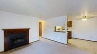 This 2-bedroom, 1-bathroom Deluxe residence is a single-floor layout designed for functional, comfortable living. The living room serves as the heart of the home, featuring plush wall-to-wall carpeting that adds warmth underfoot. A wood-mantled fireplace with a sleek black surround anchors one corner of the room, providing both a visual focal point and a cozy source of ambiance. White walls and a textured ceiling keep the space feeling bright and clean throughout.

A pass-through window connects the living area to the kitchen, promoting an open, communicative flow between the two spaces. The kitchen is equipped with warm wood-toned cabinetry, a countertop bar ledge ideal for casual dining or meal prep, and tile flooring that contrasts neatly with the carpeted living area. Under-cabinet lighting adds a practical touch to the kitchen's functionality.

A hallway visible from the living room leads to the two bedrooms and bathroom, keeping the private areas of the home clearly separated from the common living spaces. Overall, the layout balances openness with defined zones, making it a well-organized and inviting residence.