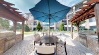 Located at 1201 Central Haven Dr in Mount Pleasant, South Carolina, this outdoor community space offers a well-appointed courtyard retreat designed for relaxation and social gathering. The centerpiece of the patio area is a round dining table surrounded by comfortable chairs, shaded by a large blue market umbrella — ideal for enjoying the South Carolina sunshine in comfort. The patio is finished with an attractive interlocking paver surface that adds texture and visual warmth to the outdoor setting. Additional seating areas are scattered throughout the courtyard, complemented by wooden pergola structures that frame the space and provide architectural character. Neatly manicured landscaping, including tall evergreen shrubs and ground-level plantings, creates a sense of privacy and greenery throughout. The surrounding multi-story brick residential buildings form a cohesive backdrop, enclosing the courtyard in a welcoming, community-oriented atmosphere. The open sky above allows for abundant natural light, making this an inviting space throughout the day. The overall ambiance is clean, modern, and thoughtfully designed to encourage outdoor living and community connection in a serene neighborhood setting.