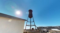 This single-floor space is defined by a standout rooftop presence anchored by a weathered, industrial-era water tower rising prominently above the surrounding roofline. The water tower, mounted on a sturdy latticed iron frame, showcases a patinated metal tank that speaks to the building's historical character and adds a strong architectural focal point to the exterior. The rooftop area features open railing systems that frame the perimeter, offering views of the surrounding neighborhood and distant hillside terrain visible on the horizon. The elevated outdoor setting benefits from abundant natural light, with the wide-open sky creating a bright, airy atmosphere throughout the day. The structural elements of the water tower — including its cross-braced support legs and tension cables — contribute a rugged, utilitarian aesthetic that contrasts with the lighter tones of the building facade. The adjacent rooftop space provides a functional platform that balances industrial heritage with practical outdoor utility. The overall ambiance of this space is one of character-rich industrial charm, where historic infrastructure meets open-air exposure, making it a visually distinctive and memorable environment.