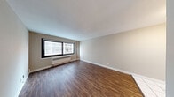 This single-floor unit offers a practical and well-organized layout that accommodates comfortable everyday living. The living area is a generously proportioned space finished with rich, dark hardwood-style flooring and neutral beige-toned walls. A large window with a black frame allows natural light to fill the room while offering views of the surrounding building exterior. A baseboard radiator provides efficient heating along the window wall.

The separate bedroom continues the same neutral color palette with soft carpeting underfoot, a ceiling fan with a light fixture, and its own window with a radiator — creating a cozy and functional retreat.

The kitchen is well-equipped with warm honey-toned wood cabinetry spanning both upper and lower sections, offering ample storage. White appliances — including a refrigerator, electric range, built-in microwave, and dishwasher — complement the light tile flooring and white countertops, giving the kitchen a clean and classic appearance.

The bathroom features patterned mosaic tile flooring, a dark wood vanity with a wall-mounted mirror, and a tub-shower combination enclosed by a striped curtain. The white walls and overhead lighting keep the space feeling bright and functional. Overall, the unit presents a clean, move-in ready environment with consistent finishes throughout.