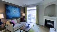 This single-floor space presents a thoughtfully designed living area that balances comfort and contemporary style. The living room serves as the focal point, anchored by a plush cream sofa accented with richly colored throw pillows in deep plum and magenta. A large-scale abstract wall art piece in vivid shades of blue commands attention on the dark feature wall, adding a bold artistic element to the space.

Natural light flows generously into the room through full-height sliding glass doors that open directly onto a private balcony, where outdoor seating is visible. Chartreuse patterned drape panels frame the doors, introducing a vibrant contrast against the neutral walls and adding visual warmth to the space.

Crown molding runs along the ceiling perimeter, lending an architectural refinement to the room. A rustic wood-and-metal coffee table sits atop a green-toned area rug, grounding the seating area and adding textural variety. The overall ambiance is contemporary yet inviting, with a carefully curated mix of bold color accents, natural textures, and functional furnishings that create a cohesive and welcoming environment.