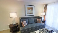 This single-floor space presents a thoughtfully arranged living area that combines comfort and style. The living room serves as the focal point, anchored by a substantial blue-gray upholstered sofa accented with decorative throw pillows in complementary rust and tan tones. Framed wall art above the sofa adds a refined touch, depicting a simple still-life composition that reinforces the room's warm, neutral color palette. Flanking the seating area are two matching table lamps with square shades, providing balanced ambient lighting and contributing to the room's symmetrical aesthetic. A dark-finished side table sits adjacent to the sofa, offering functional surface space. Drapery panels in warm rust tones frame what appears to be a window, allowing natural light into the space. The carpeted flooring adds warmth underfoot and ties the room together. Additional seating with bold patterned upholstery is partially visible, suggesting a versatile and well-furnished layout. Overall, the space presents a cohesive, inviting atmosphere with carefully selected furnishings and a consistent warm-toned design scheme throughout.